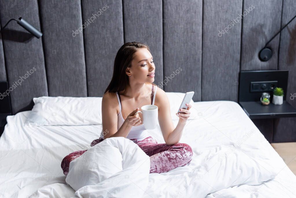 attractive young woman using smartphone and holding cup of coffee in bed 