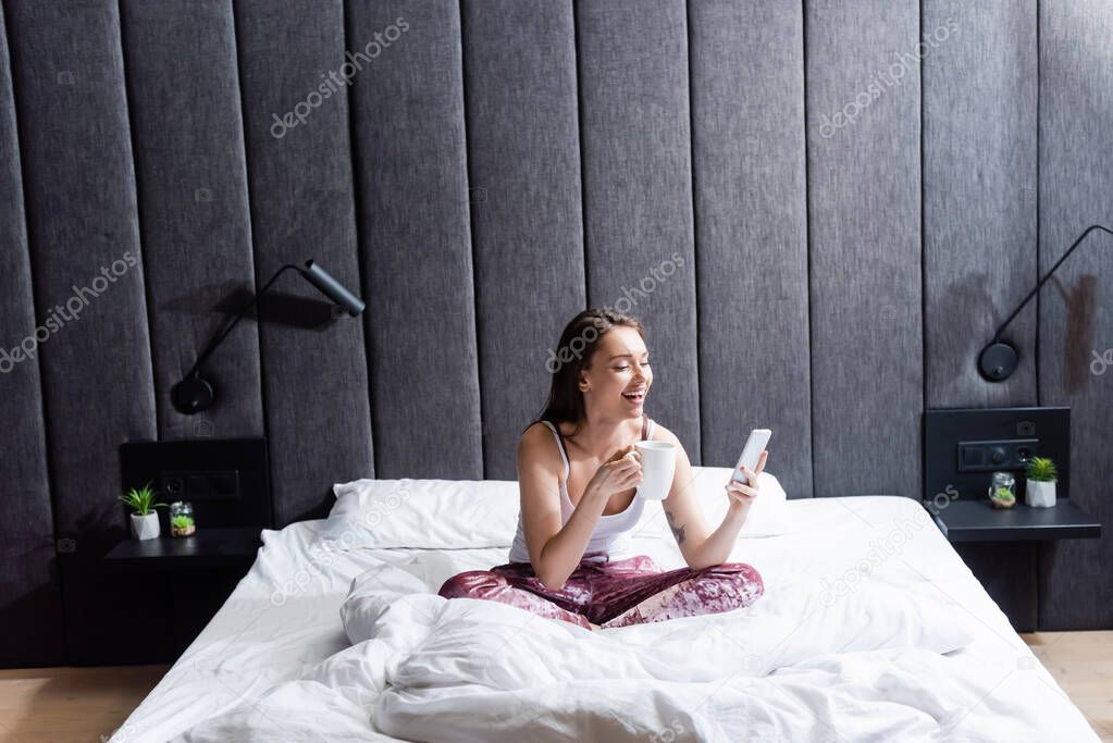 happy young woman using smartphone and holding cup of coffee in bed 