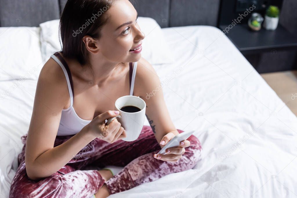 happy young woman holding smartphone and cup of coffee in bed 