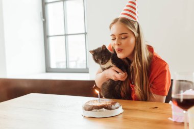 Doğum günü pastasında mum üfleyen ve kollarında sevimli bir kedi tutan seçici bir kız. 