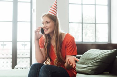 Parti şapkası takmış neşeli bir kız evde akıllı telefondan konuşuyor. 