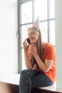 cheerful girl in party cap sitting on window sill and talking on smartphone clipart