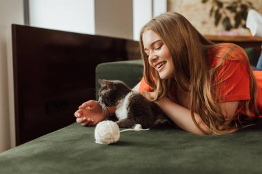 Neşeli kız kanepede iplik yumağının yanında tatlı kedi oynuyor.