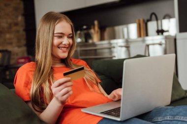 Bilgisayarın yanında kredi kartı tutan mutlu genç bir kadının seçici odak noktası 