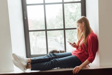 Genç bir kadın pencerenin kenarında kedisiyle oturuyor ve akıllı telefon kullanıyor. 