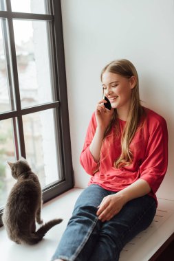 Mutlu kadın pencerenin pervazında kedisiyle oturuyor ve akıllı telefonla konuşuyor. 