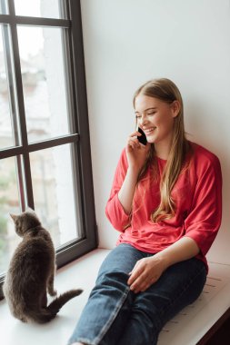 happy woman sitting on window sill with cat and talking on smartphone  clipart
