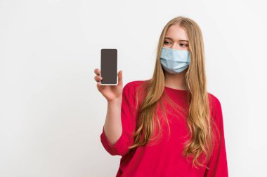 Tıbbi maskeli kadın beyaz ekranlı akıllı telefona bakıyor. 