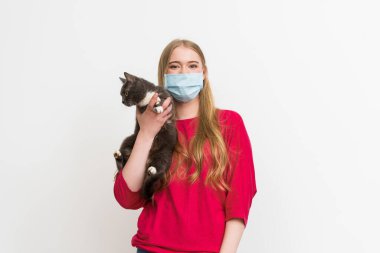 Tıbbi maskeli genç bir kadın kucağında sevimli bir kedi ve beyaz kameraya bakıyor. 