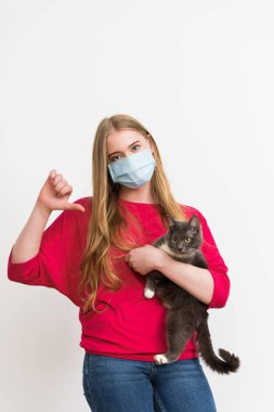Tıbbi maskeli genç bir kadın kucağında sevimli bir kedi ve baş parmağını beyazın üzerinde izole bir şekilde gösteriyor. 