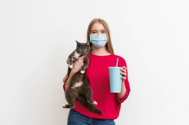 Tıbbi maskeli genç bir kadın kucağında sevimli bir kedi ve tek kullanımlık gazoz dolu beyaz bir bardak tutuyor. 