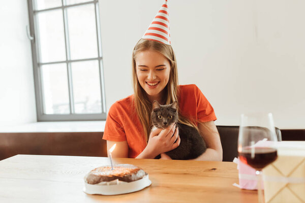selective focus of happy girl in party cap looking at birthday cake and holding in arms cat 