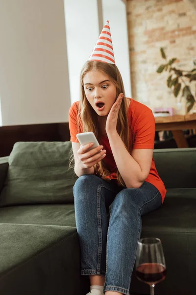 selective focus of surprised girl in party cap sitting on sofa and looking at smartphone