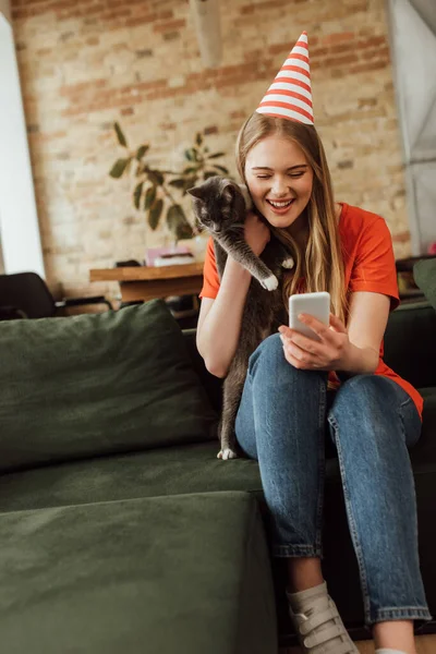 Cheerful Young Woman Party Cap Holding Cat Using Smartphone — Stock Photo, Image