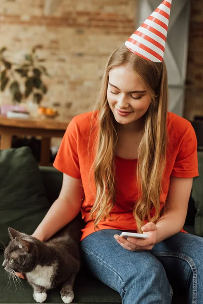 Wesoła Młoda Kobieta Party Cap Dotykając Ładny Kot Trzymając Smartfona — Zdjęcie stockowe