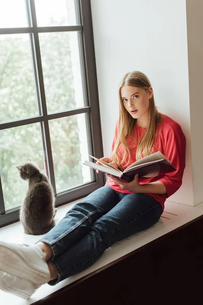 Vacker Flicka Håller Bok Och Sitter Fönsterbräda Nära Söt Katt — Stockfoto