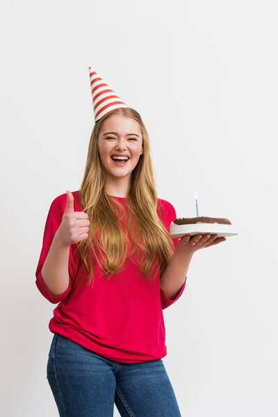 cheerful girl in party cap showing thumb up and holding birthday cake isolated on white 