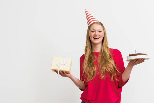 веселая девушка в шапочке для вечеринок с праздничным тортом и подарком, изолированным на белом
 