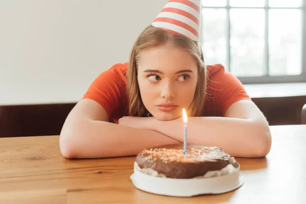 selective focus of sad woman in party cap looking away near burning candle on delicious birthday cake