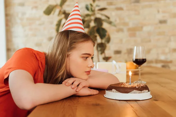 beautiful girl in party cap looking at tasty birthday cake near wine glass and presents on table