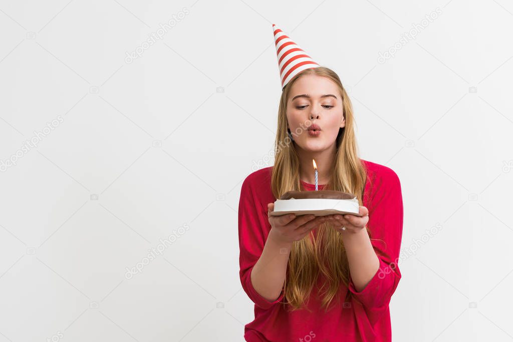 pretty girl in party cap blowing out candle on birthday cake isolated on white 