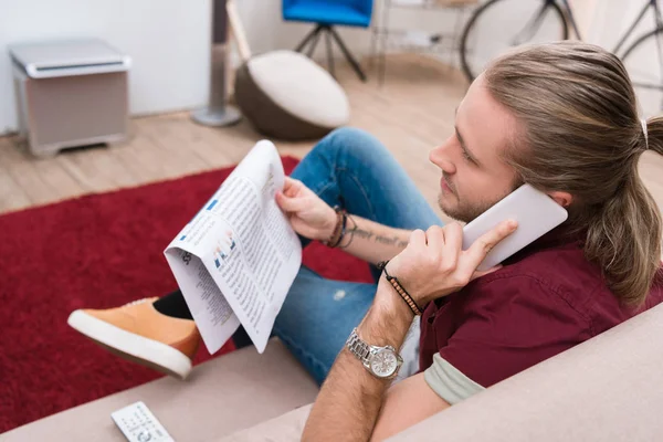 Hombre guapo leyendo el periódico y hablando en el teléfono inteligente en casa - foto de stock