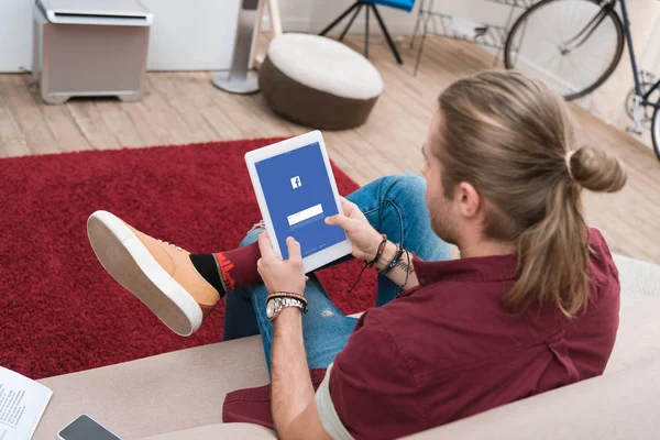 Homme assis sur le canapé tout en utilisant une tablette numérique avec appareil facebook — Photo de stock