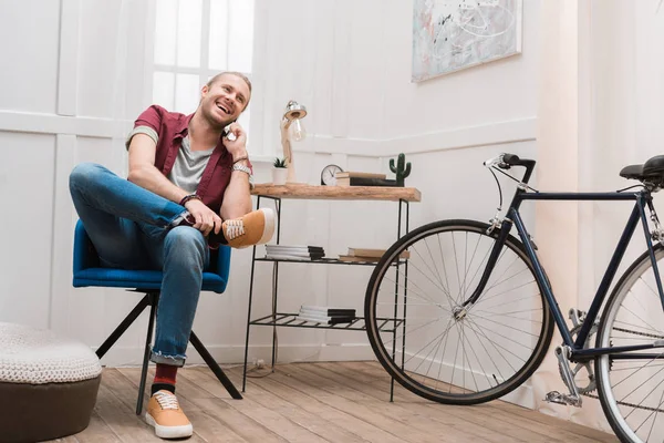 Homme souriant parlant sur smartphone tout en étant assis sur une chaise à la maison avec vélo — Photo de stock