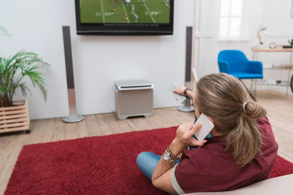 Visão traseira do homem falando no smartphone enquanto assiste jogo de futebol em casa — Fotografia de Stock