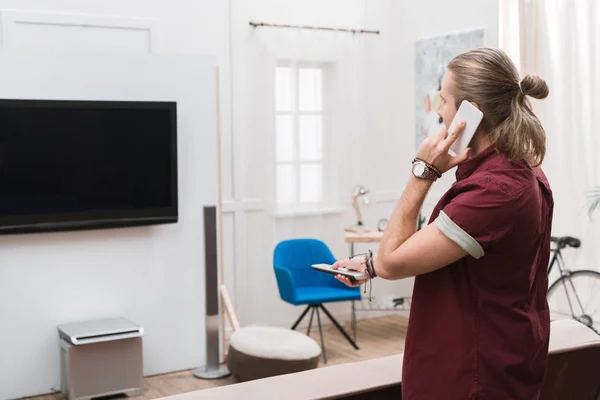 Schöner Mann mit Fernbedienung, der auf dem Smartphone spricht und zu Hause fernsieht — Stockfoto