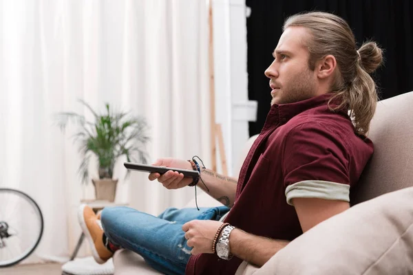 Handsome concentrated man with remote control watching tv at home — Stock Photo