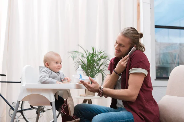 Padre alimentando a su pequeño hijo con comida para bebés mientras habla en el teléfono inteligente - foto de stock