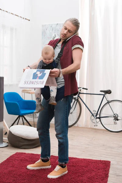 Pai de pé com o filho na funda do bebê, segurando jornal e falando por smartphone — Fotografia de Stock