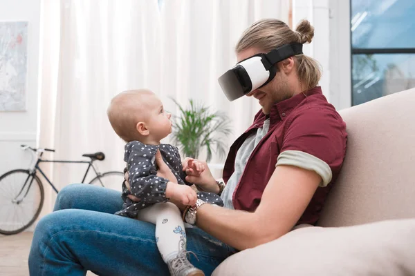 Улыбающийся отец сидит со своей маленькой дочерью и использует гарнитуру виртуальной реальности — стоковое фото