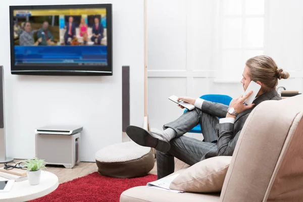 Businessman talking by smartphone and changing TV channels at home — Stock Photo