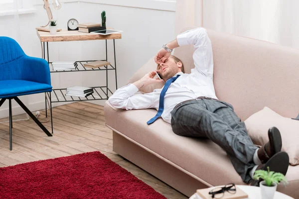 Tired businessman lying on a sofa at home and talking by smartphone — Stock Photo