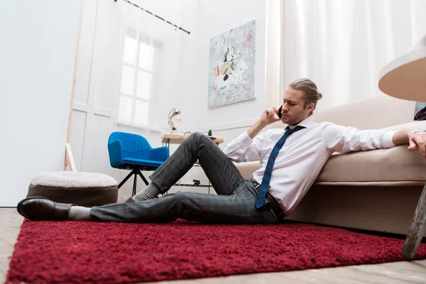 Бизнесмен сидит дома на красной ковровой дорожке и разговаривает по смартфону — стоковое фото