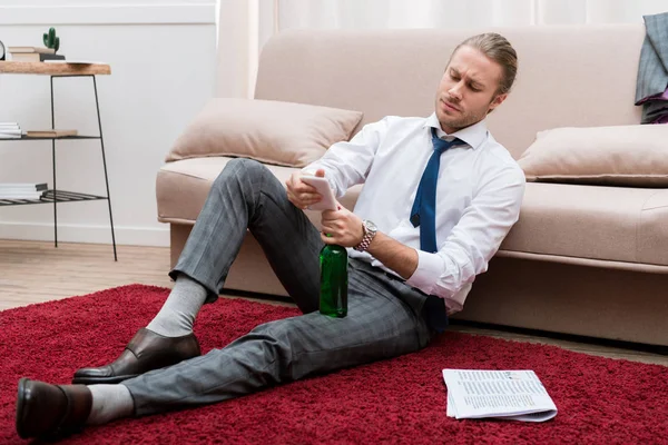 Bonito homem sentado em um andar na sala de estar com smartphone e garrafa de cerveja — Fotografia de Stock