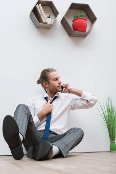 Businessman sitting on a wooden floor, talking by smartphone and loosening tie — Stock Photo