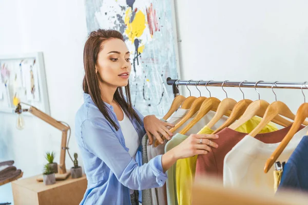 Beautiful designer looking at clothes on hangers — Stock Photo