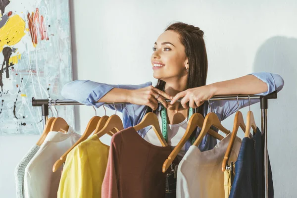 Smiling seamstress leaning on rack with hangers and clothes — Stock Photo