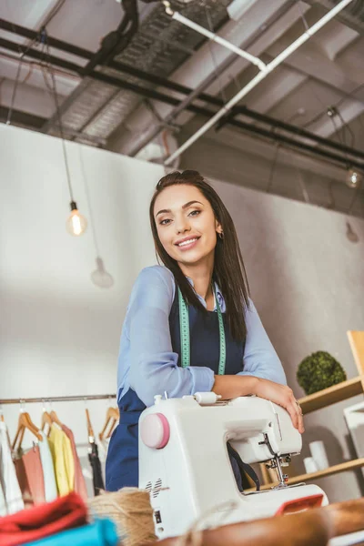 Smiling seamstress leaning on sewing machine and looking at camera — Stock Photo