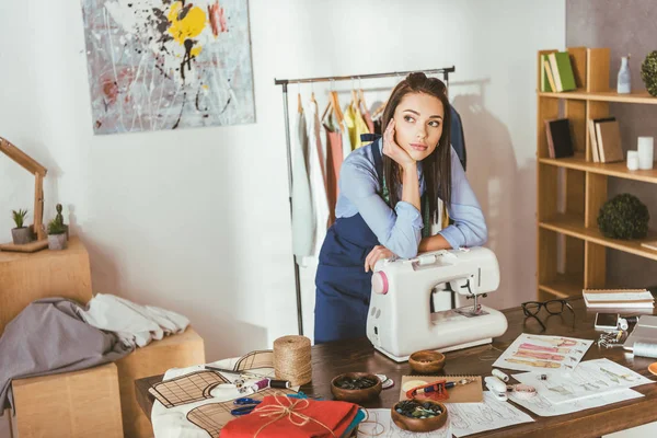 Atractiva costurera apoyada en la máquina de coser y mirando hacia otro lado - foto de stock