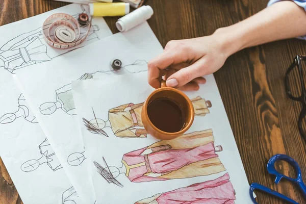 Cropped image of seamstress holding cup of tea above sketches — Stock Photo
