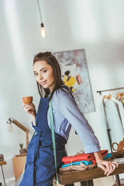 Atractiva costurera apoyada en la mesa, sosteniendo la taza de té y mirando a la cámara - foto de stock