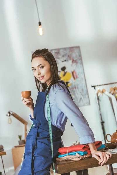 Attraktive Näherin lehnt am Tisch, hält eine Tasse Tee in der Hand und blickt in die Kamera — Stockfoto