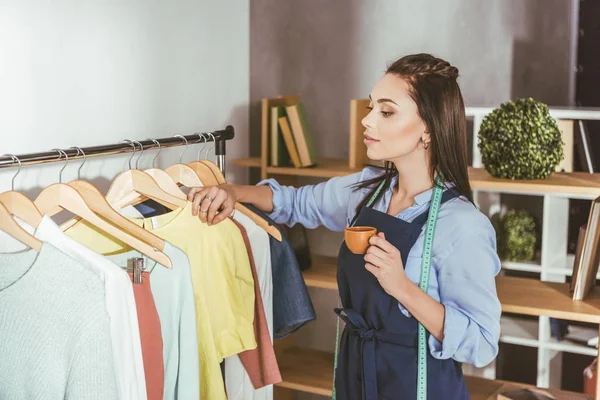 Seamstress looking at clothes on hangers and holding cup of tea — Stock Photo