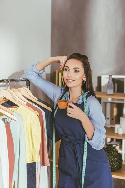 Glückliche Näherin lehnt sich an Gestell mit Kleidern auf Kleiderbügel — Stockfoto