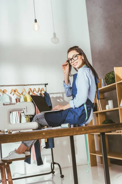 Costurera feliz sentado en la mesa con el ordenador portátil y mirando a la cámara - foto de stock