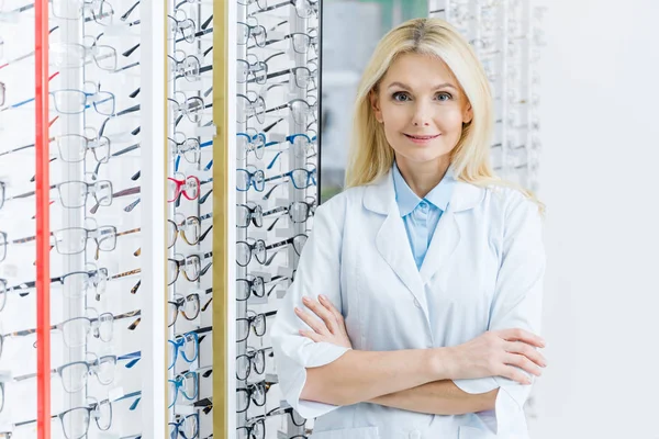 Blonde female ophthalmologist standing in optics with glasses on shelves — Stock Photo